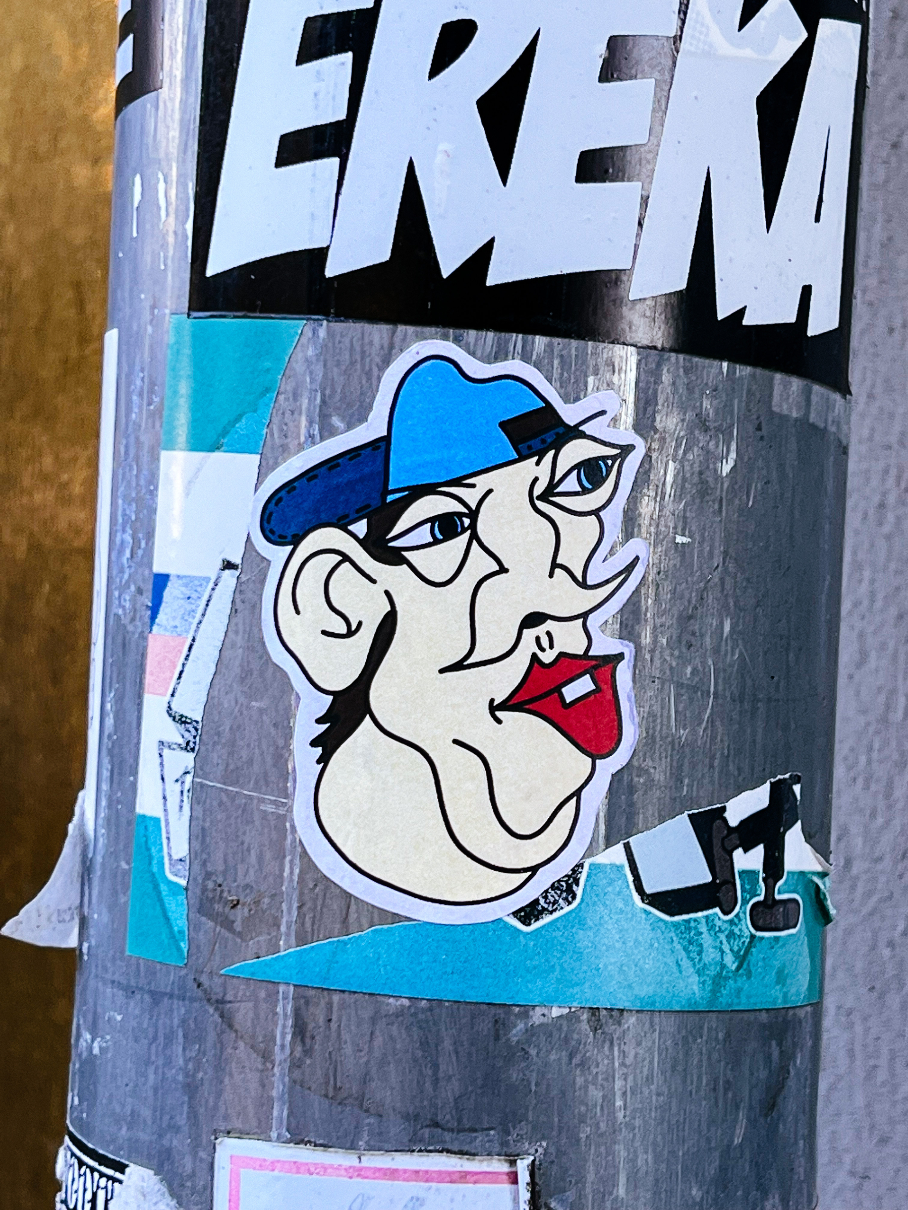 Sticker with a drawing of a strange looking face, red lips, and and blue cap. 