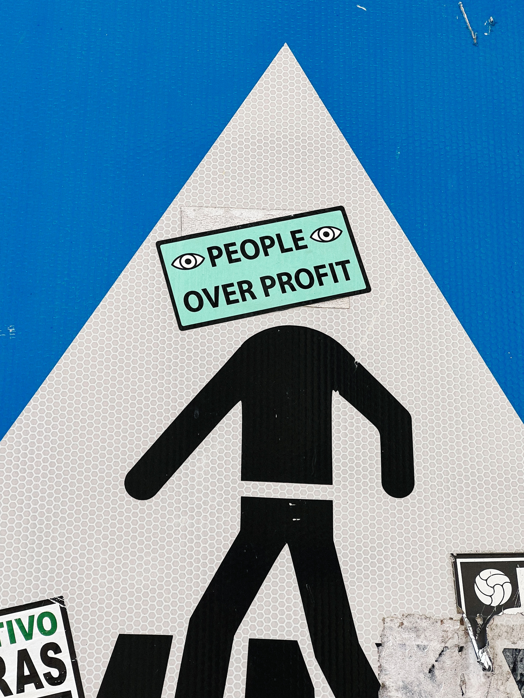 A crosswalk street sign where the head of the person crossing the street has been replaced with a sticker that says “people over profit”. 