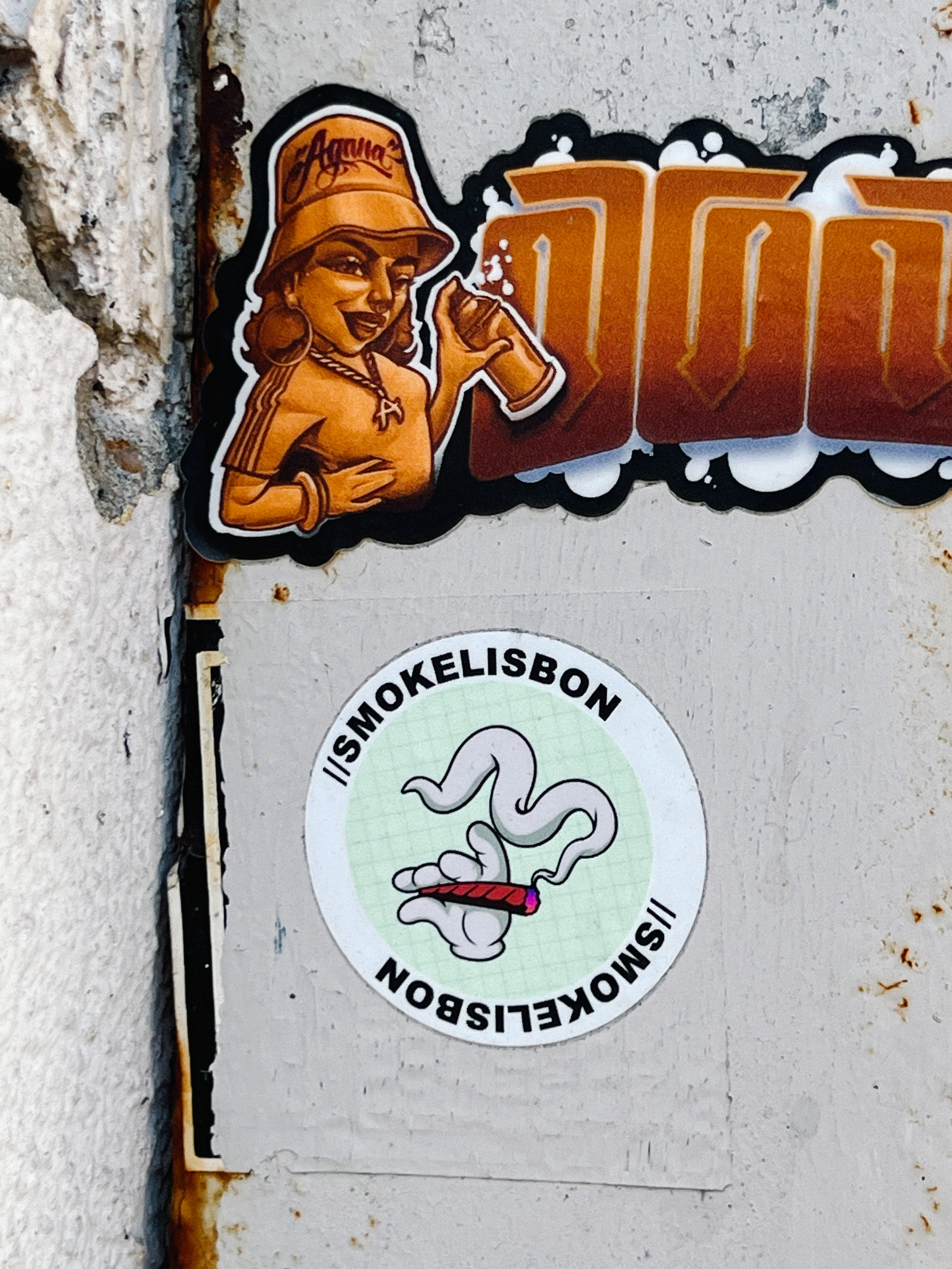 Two stickers. One of a graffiti artist, can in hand, and another one with a hand holding a joint, and “smokelisbon” written on the border. 