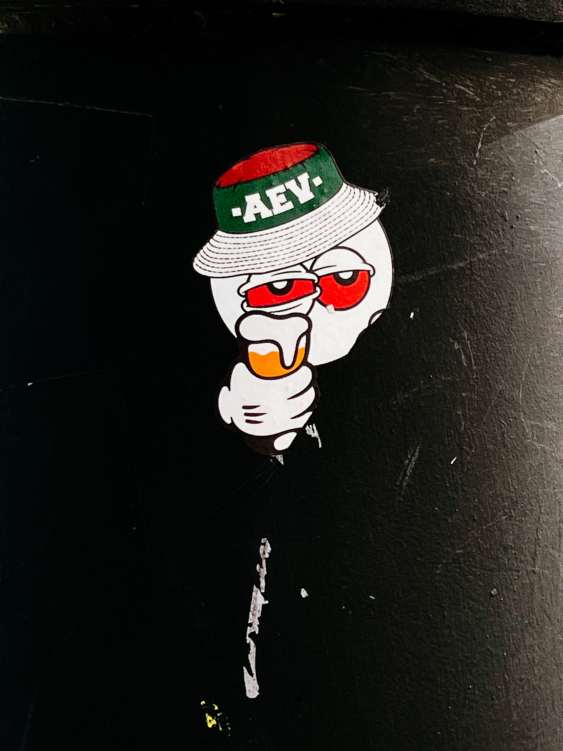 Sticker with a white ball as a face, red eyed, wearing a bucket hat with “-AEV-“ written on it. A Mickey Mouse-like hand holds a beer. 
