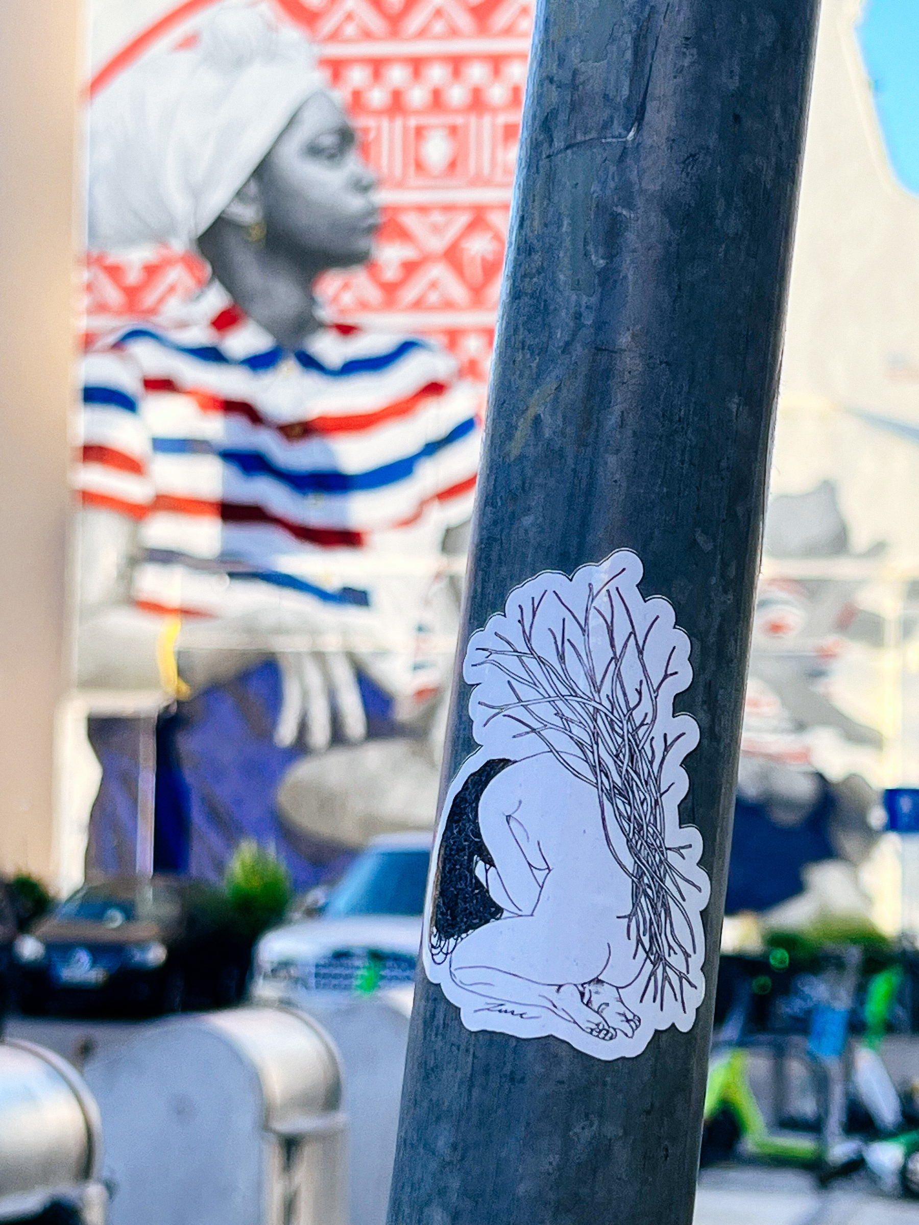 Sticker on a post. A woman, curled up, with branches on her back. In the background a graffiti of a woman, out of focus. 