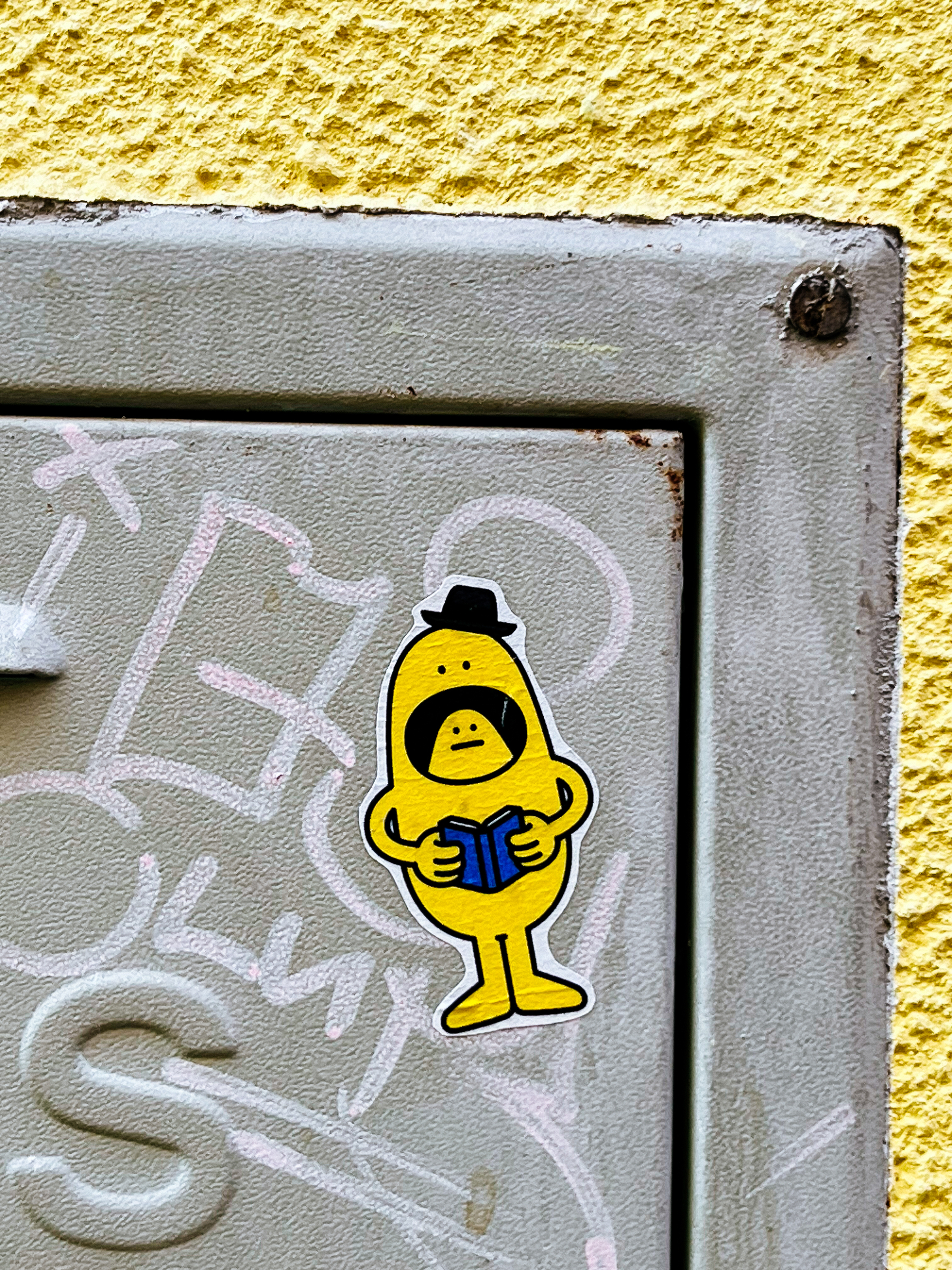 Sticker of a yellow character reading a blue book, and wearing a hat. His mouth is open, and there&rsquo;s a face inside it, like if another character was hiding inside. 