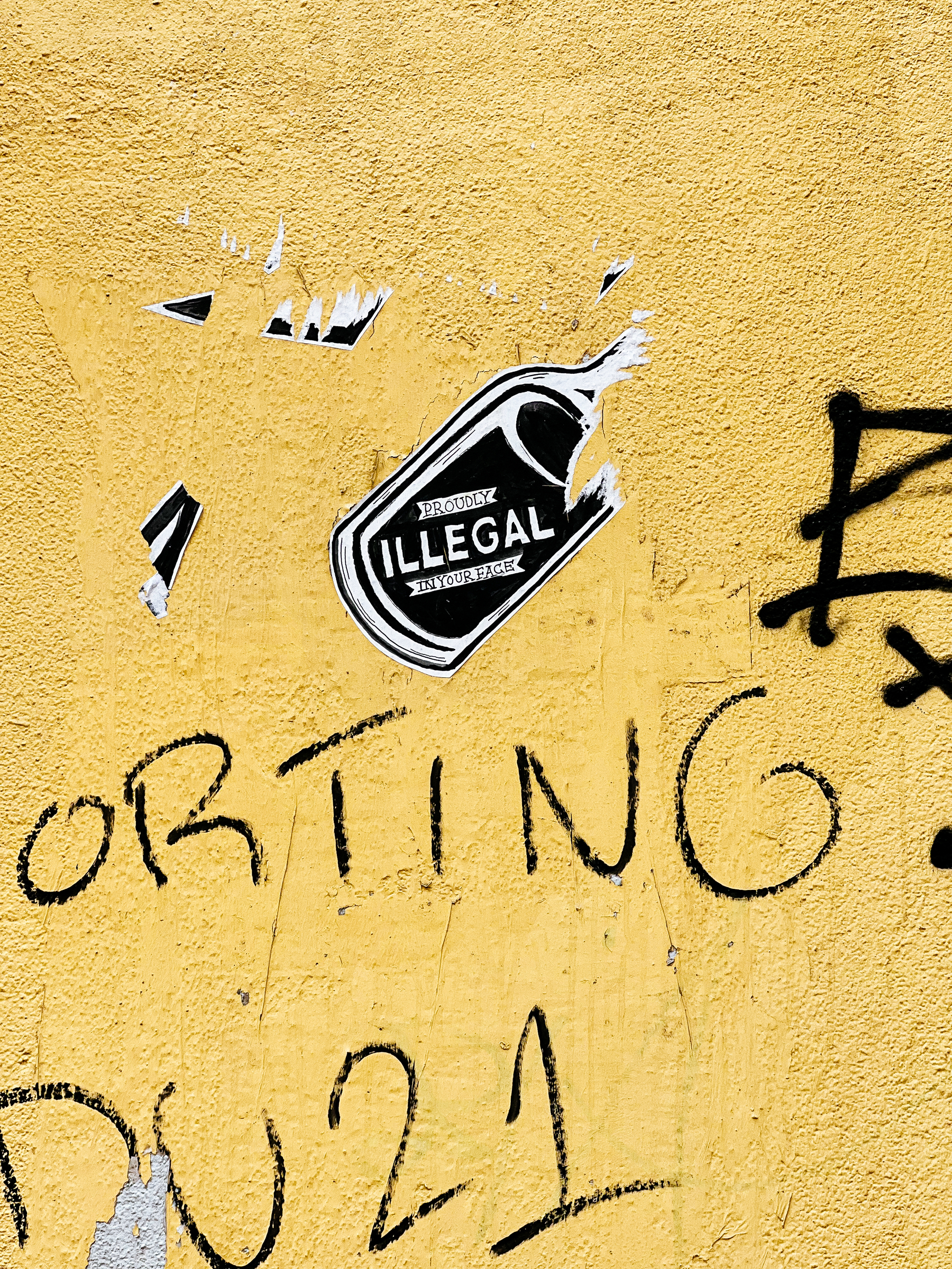 What’s left of a bottle sticker with the words “proudly illegal in your face”. Yellow wall. 