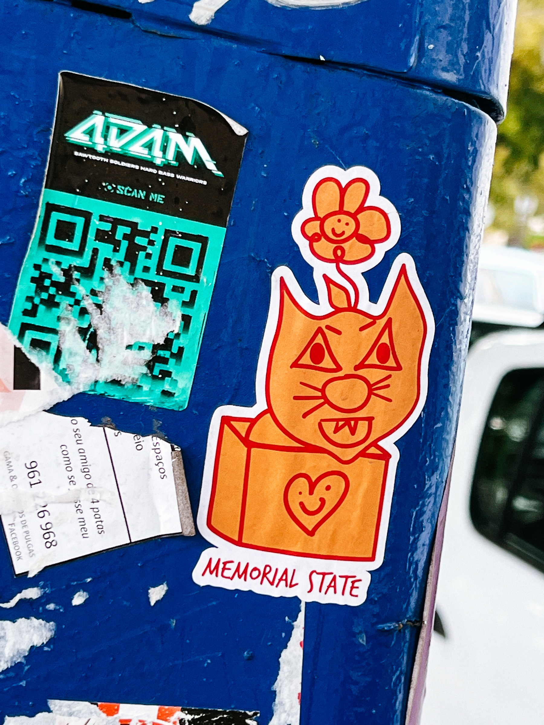 A sticker with a drawing of a cat. The cat has a flower on his head. The cat is inside a box. The box has a heart drawn on the side. 