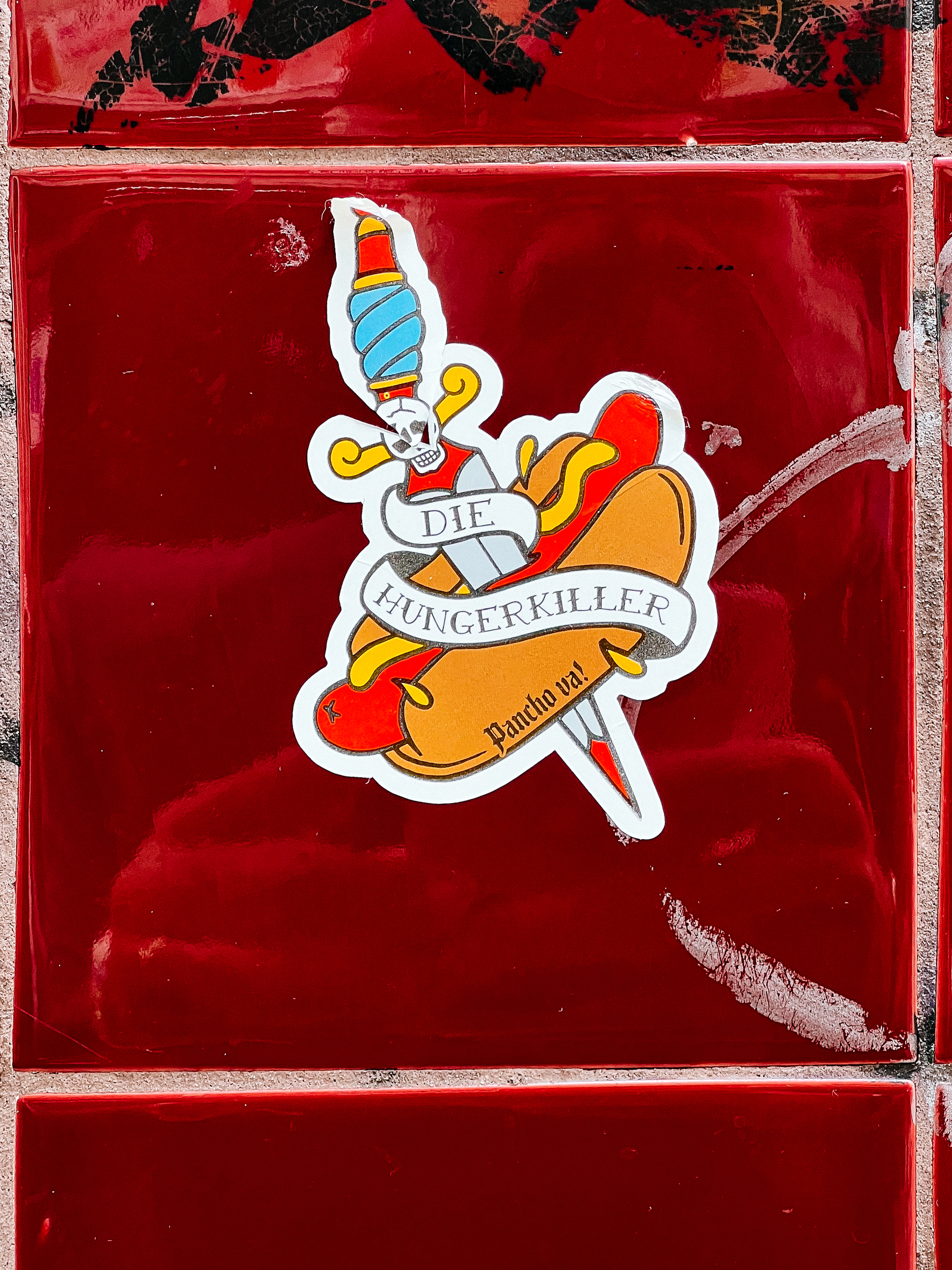 A sticker in classic American tattoo style, with a knife stuck on a hot dog. The words “die hungerkiller” in a banner. 