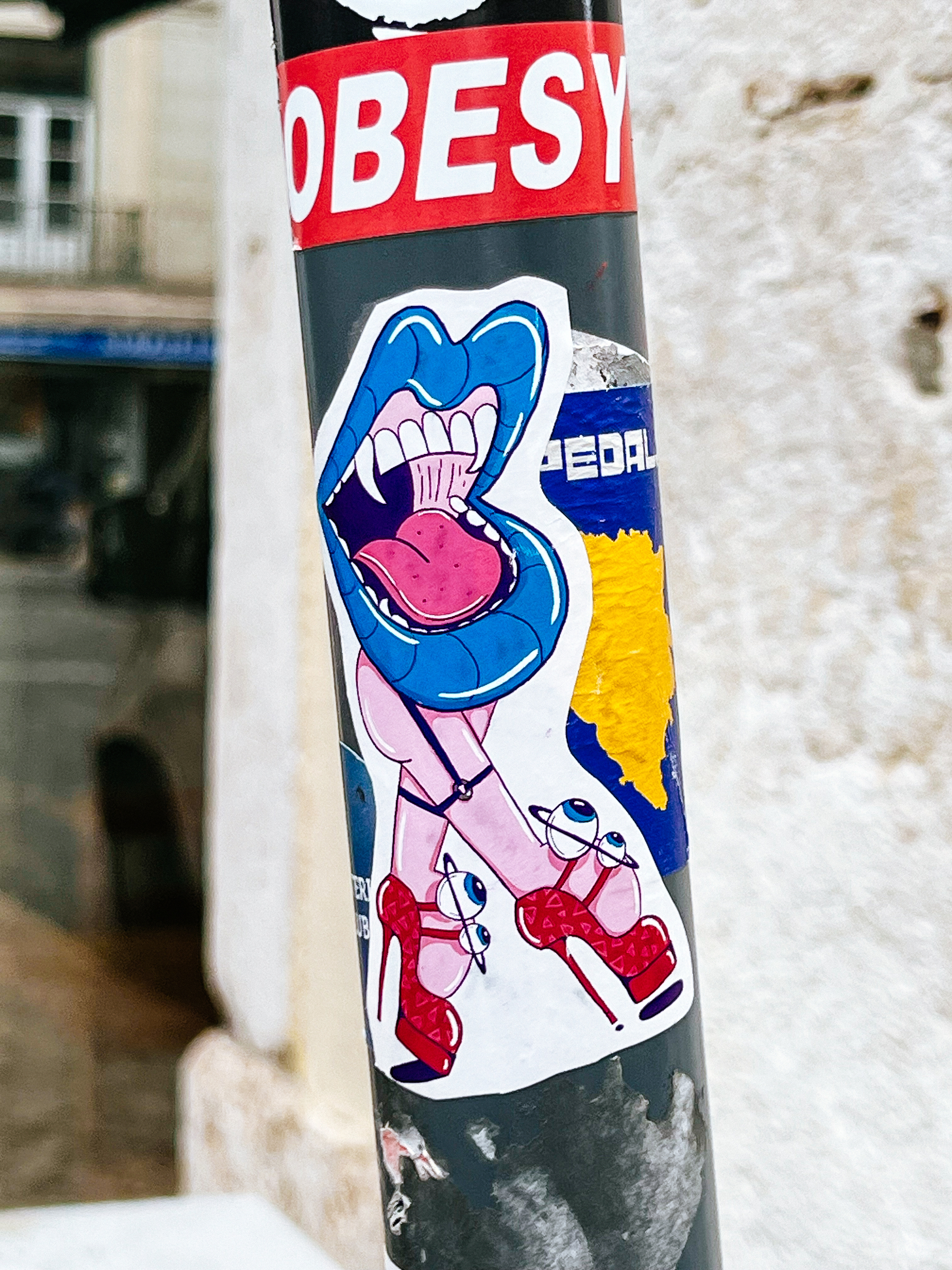 Sticker with cartoon style design. A mouth with fangs, and blue lips. Beneath it a pair of legs, wearing red high heel shoes. The shoes have eyes on the straps. 