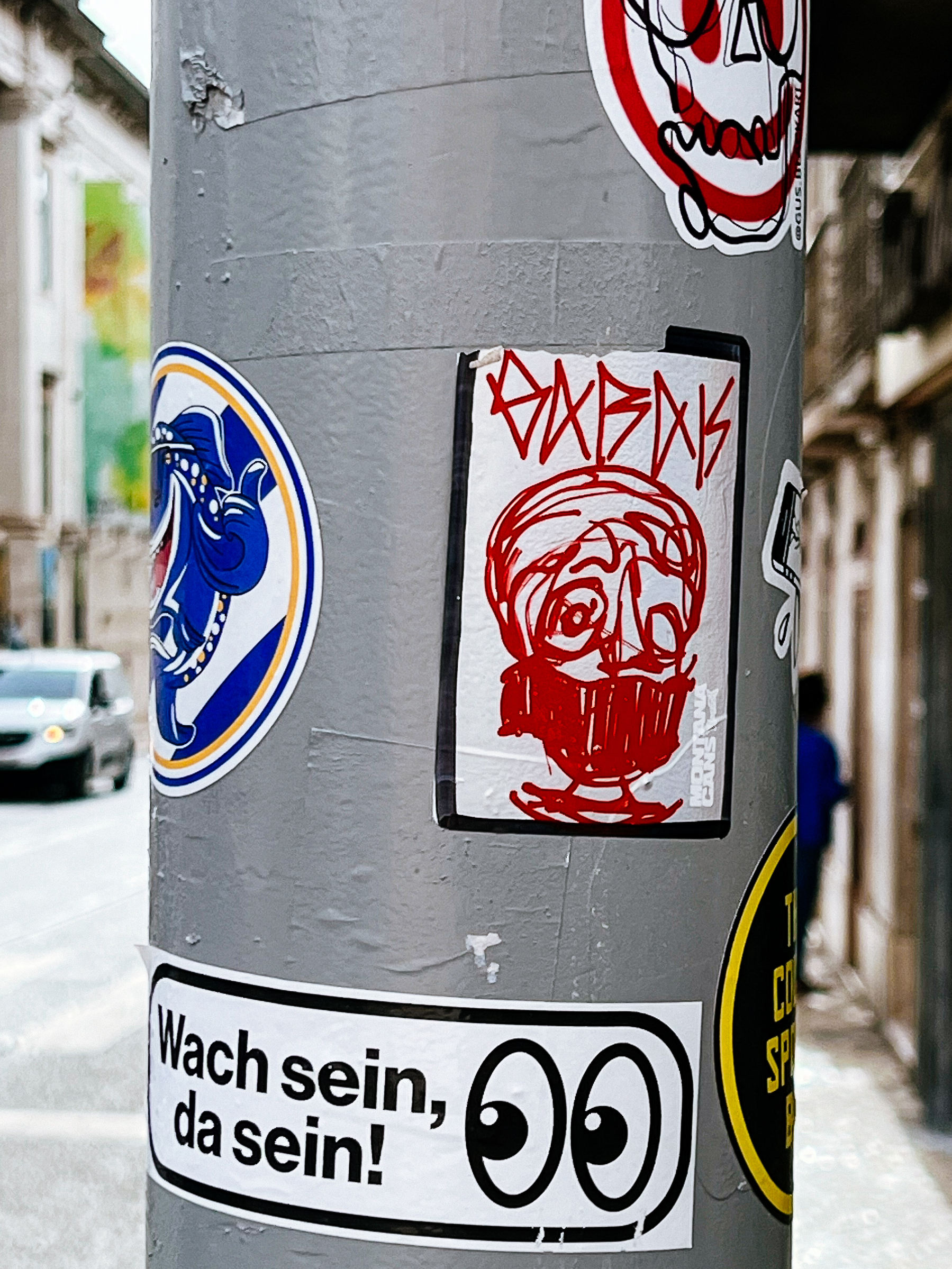 Stickers on a utilities pole. One has a crudely drawn face, and another a pair of eyes, emoji like, and “wach sein, da sein”. 