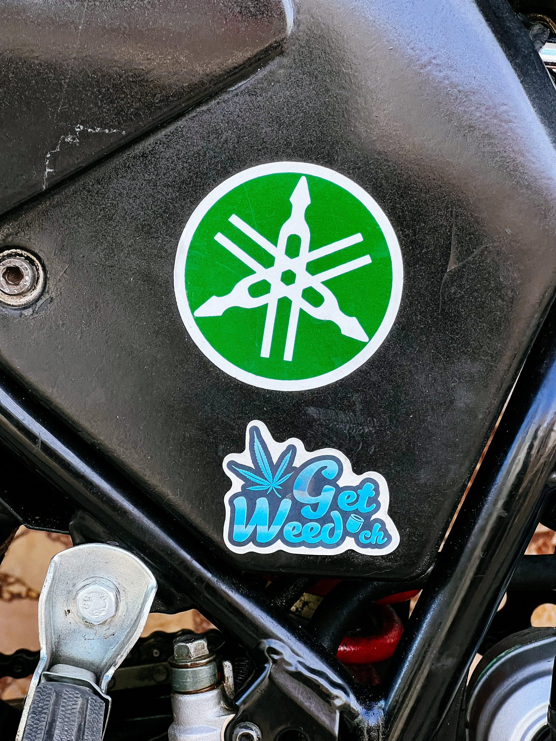 Two stickers on a motorcycle. One is the Yamaha logo, the other says “Get Weed”. 